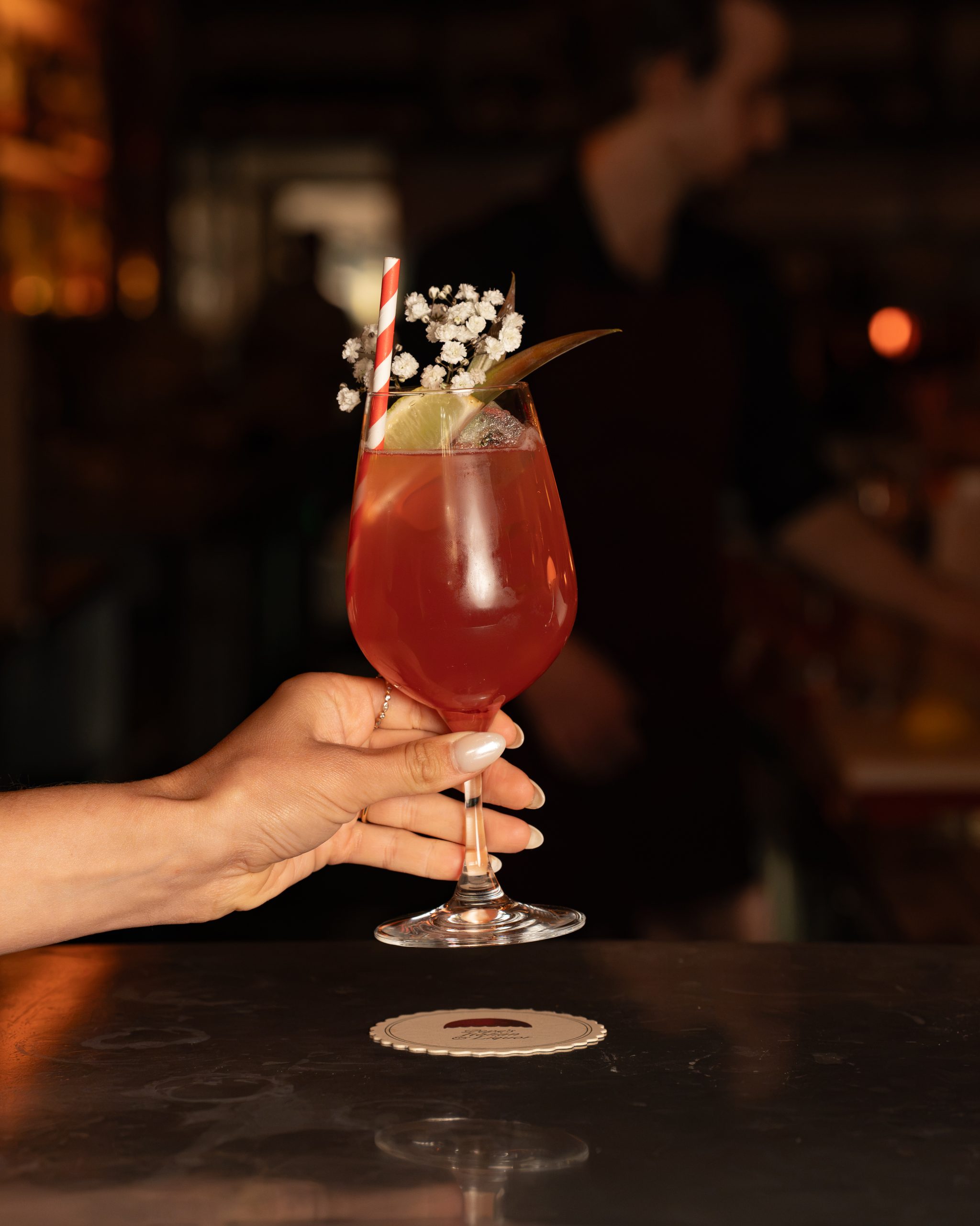 A red cocktail in a wine glass, garnished with flowers and a striped straw, at the bar